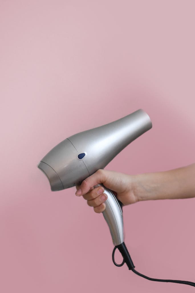 Close-up of a hand holding a silver hair dryer on a pink background.